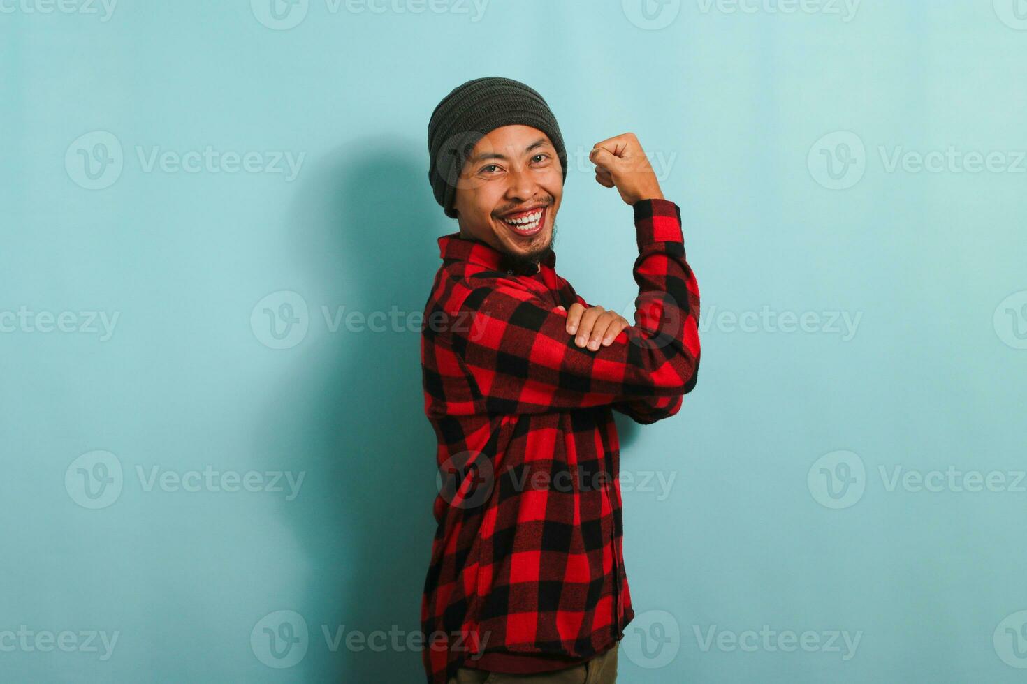 Excited Young Asian man with beanie hat and red plaid flannel shirt making a strong gesture, showing strength by flexing his arms and muscles, isolated on a blue background photo