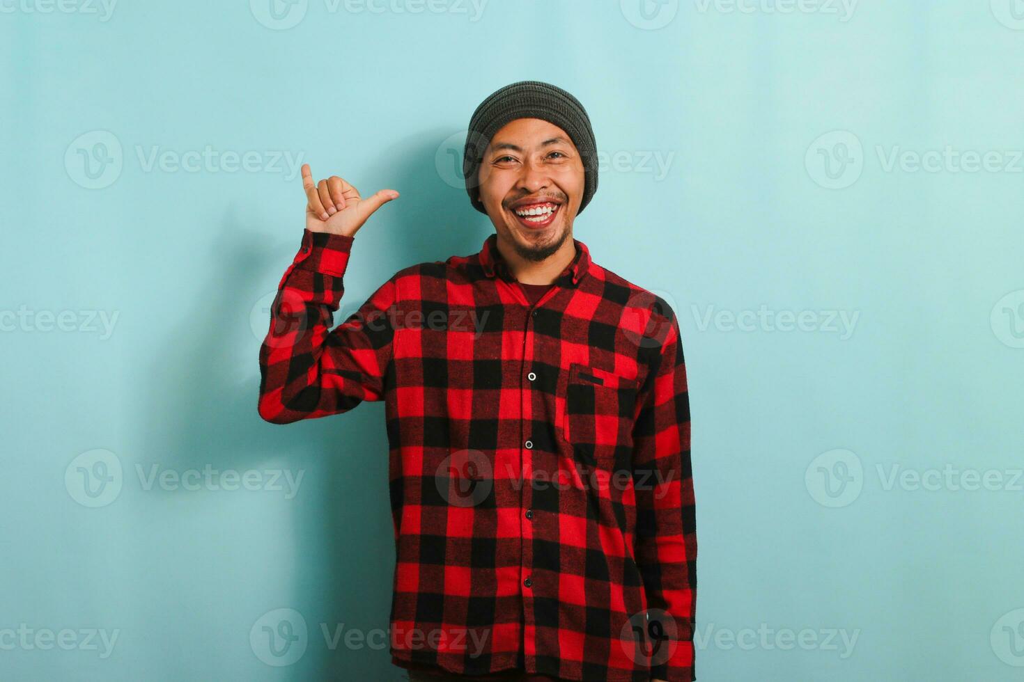 Happy young Asian man with a beanie hat and red plaid flannel shirt showing the rock and roll gesture with his fingers, expressing love for heavy metal music, isolated on a blue background photo