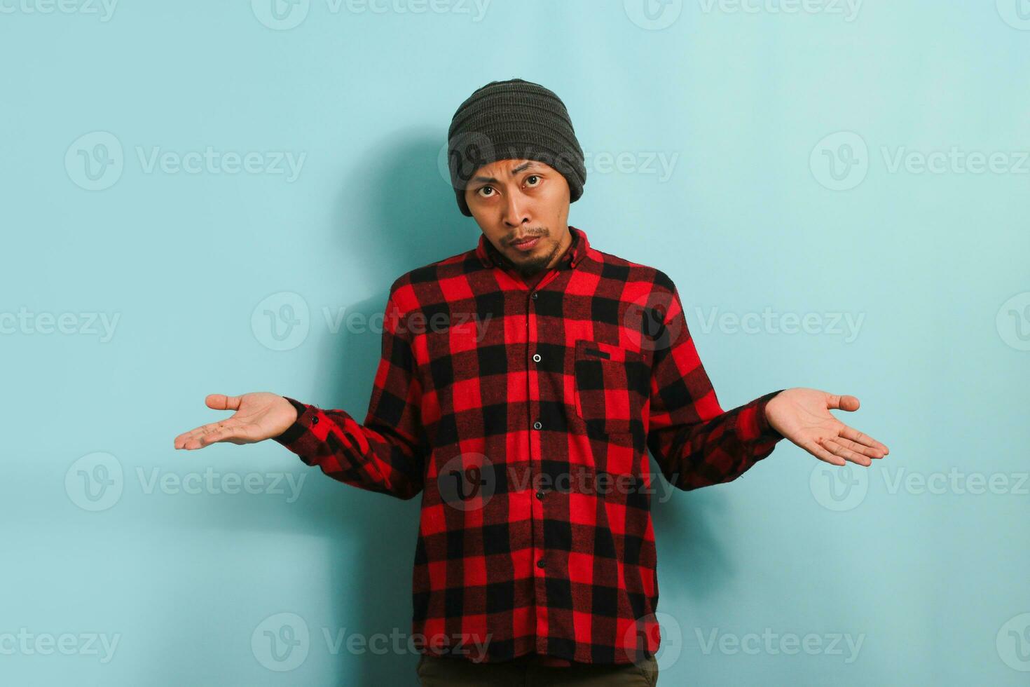A confused young Asian man with a beanie hat and a red plaid flannel shirt is shrugging his shoulders and looking at the camera with a puzzled expression, isolated on a blue background. photo