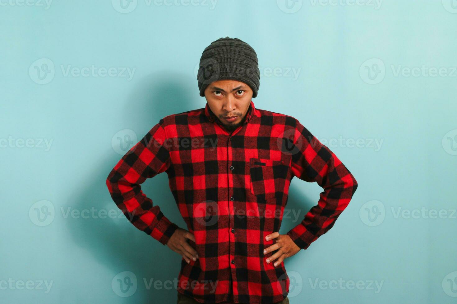 A furious young Asian man with a beanie hat and a red plaid flannel shirt stands with arms akimbo, staring directly at the camera, feeling upset. He is isolated on a blue background photo