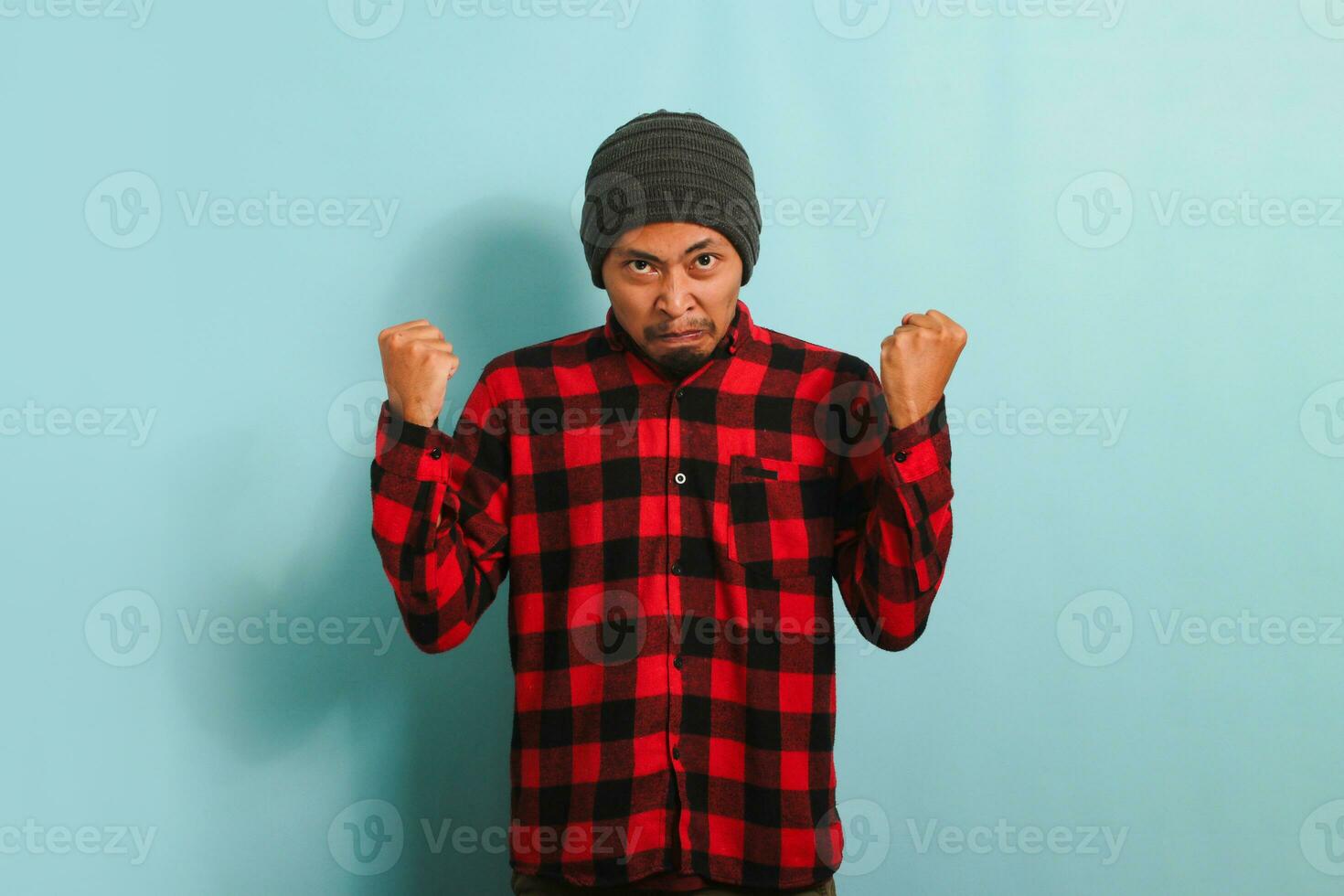 un furioso joven asiático hombre con un gorro sombrero y un rojo tartán franela camisa levanta su puño y mira furiosamente a el cámara, mostrando frustración. él es aislado en un azul antecedentes foto