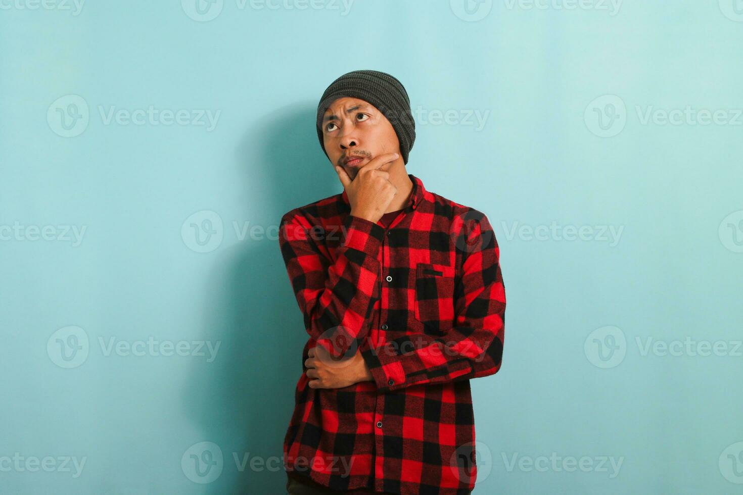 Thoughtful young Asian man with a beanie hat and a red plaid flannel shirt keeps his hand on his chin, looking aside at a copy space with a serious expression while standing against a blue background photo