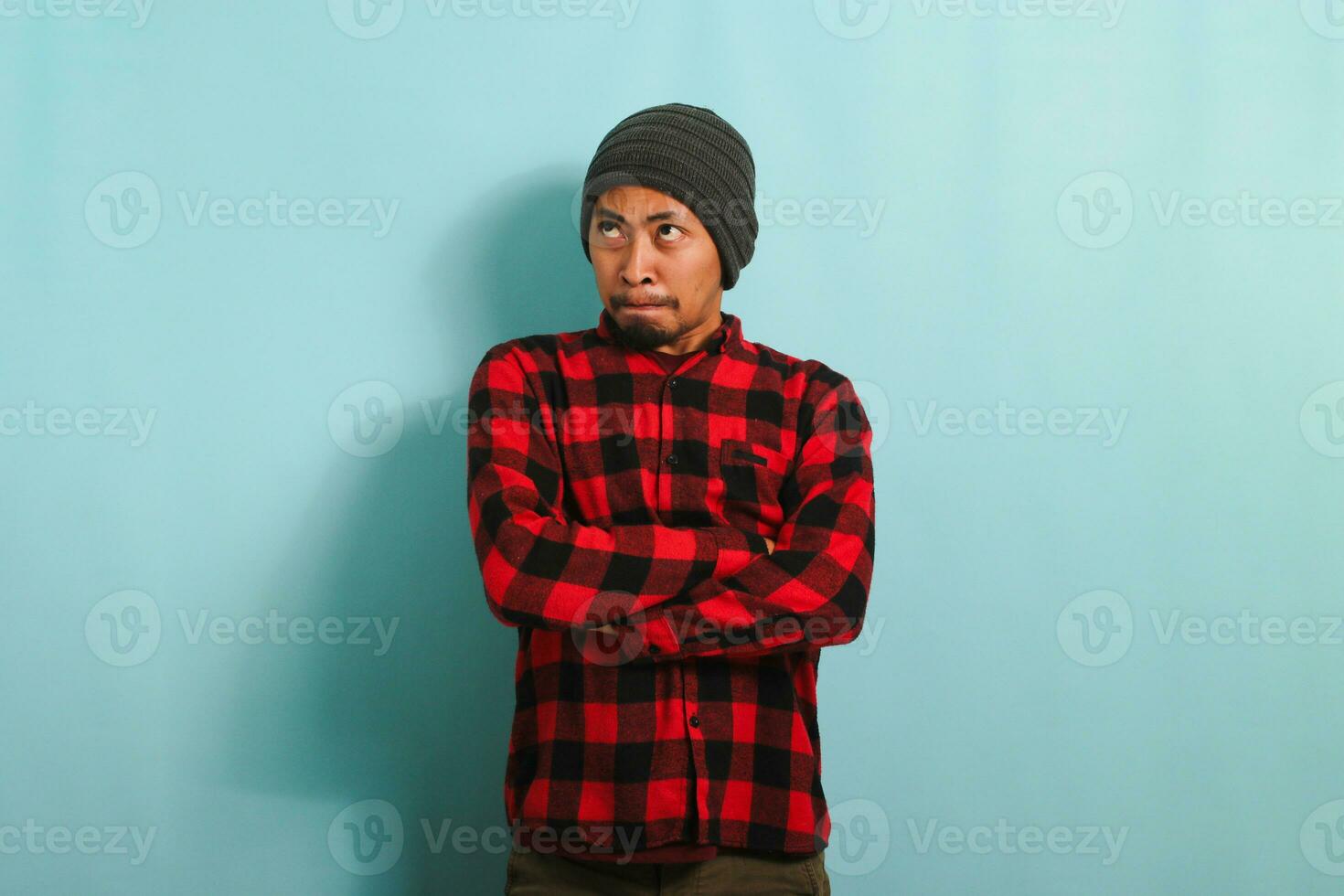 An angry young Asian man with a beanie hat and a red plaid flannel shirt keeps his arms folded, waiting for explanations with an annoyed and disapproving expression, isolated on a blue background photo