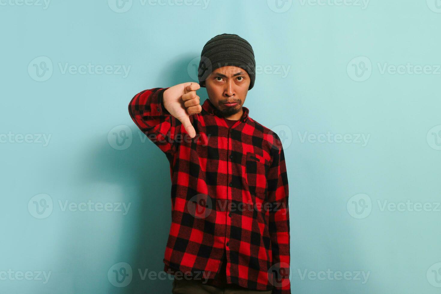 Annoyed Young Asian man with beanie hat and red plaid flannel shirt showing a thumbs-down gesture, giving a bad review, looking disappointed, frowning, and feeling sad, isolated on blue background photo