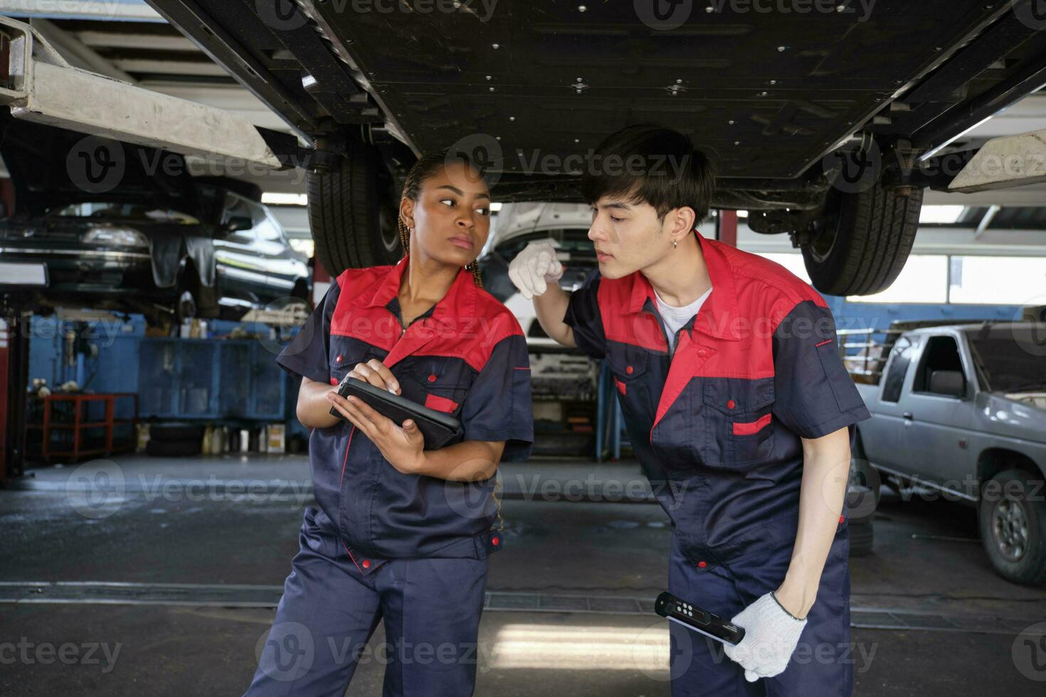 Two professional automotive worker partners discuss mechanical check undercarriage of lifted electric car, fix and repair at maintenance garage, and expert service business works industry occupation. photo