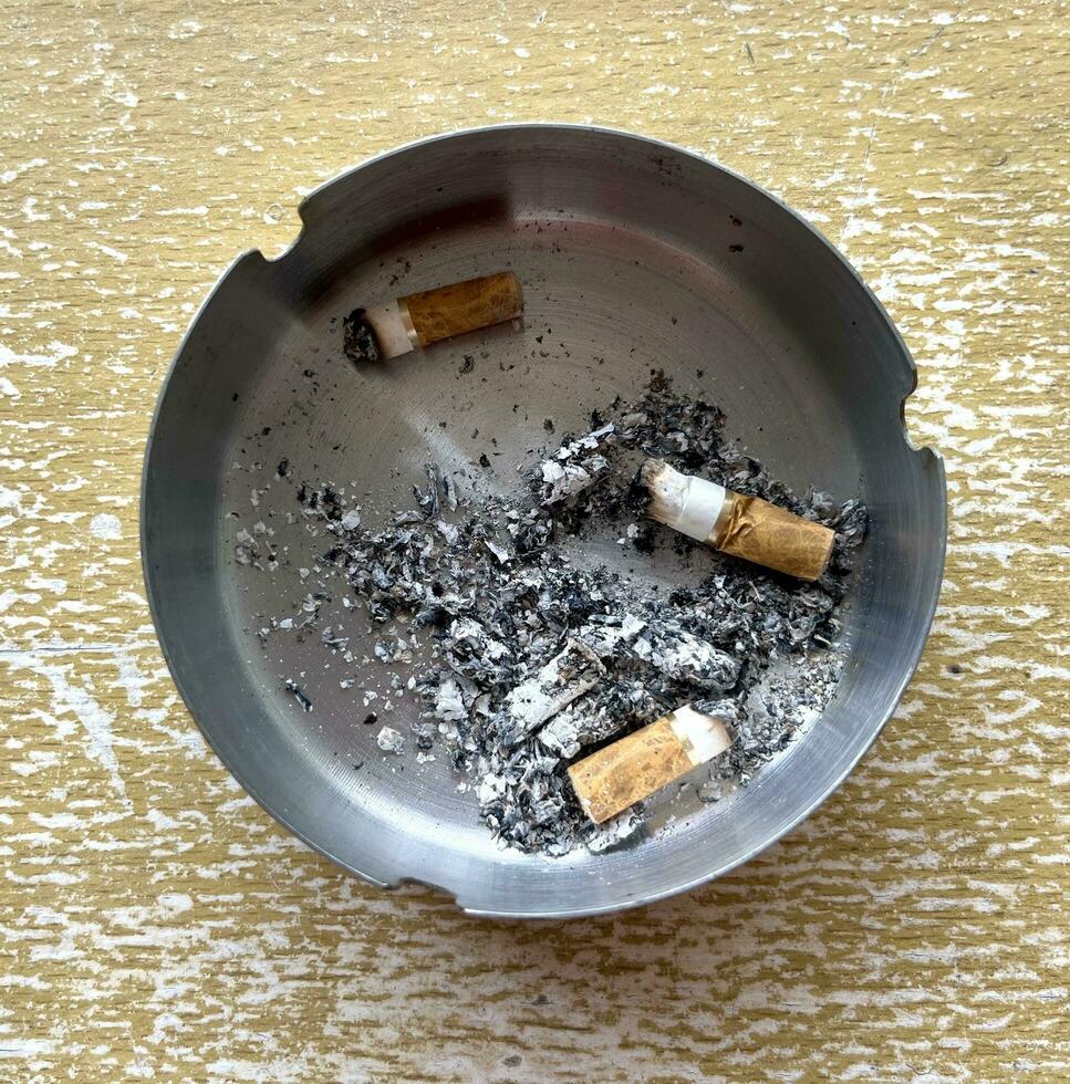 Three finished cigarette with ashes around it inside silver stainless steel ashtray isolated on wooden table background. Top view smoking equipment object photography. photo