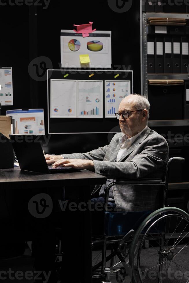 Senior businessman executive in wheelchair at office desk checking quarterly company results analysis data on laptop. Accountancy workplace filled with paperwork folders on cabinet shelves photo