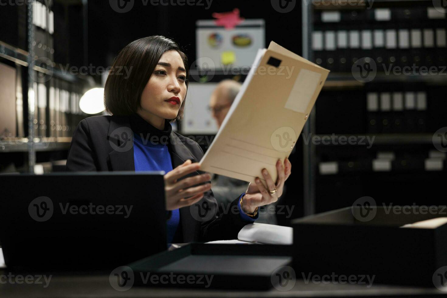 Asian private detective in agency file cabinet room reading criminal case file details. Investigator woman surrounded by criminology document folders on office repository cabinet shelves photo