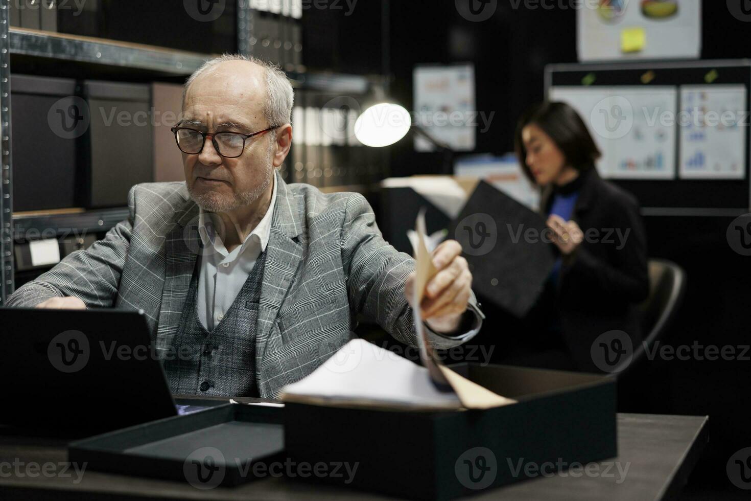 Elderly private investigator flicking through criminal case details paperwork before starting investigation. Senior detective in archive room office filled with criminology folder files photo