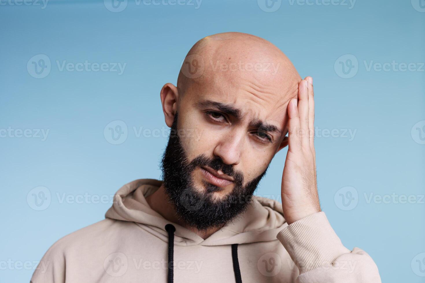 Arab man suffering from headache, holding hand on head in pain portrait. Young adult bald bearded man having migraine while looking at camera with exhausted facial expression photo