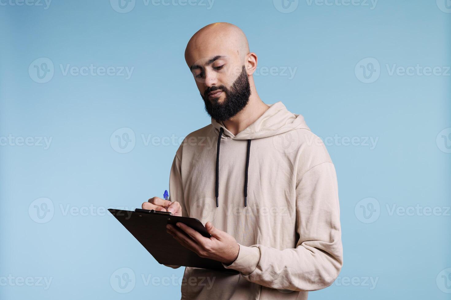 Young arab man writing in clipboard with concentrated facial expression. Focused arabian bald bearded person making list and taking notes while standing on blue background photo