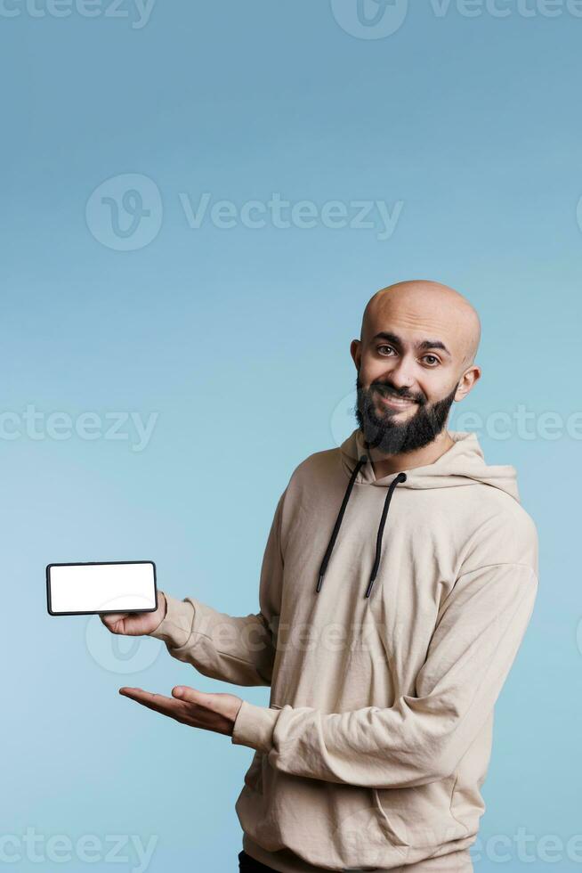 Smiling cheerful arab man showing smartphone with blank white screen for marketing mock up portrait. Carefree advertiser holding mobile phone with empty display for app promotion and looking at camera photo