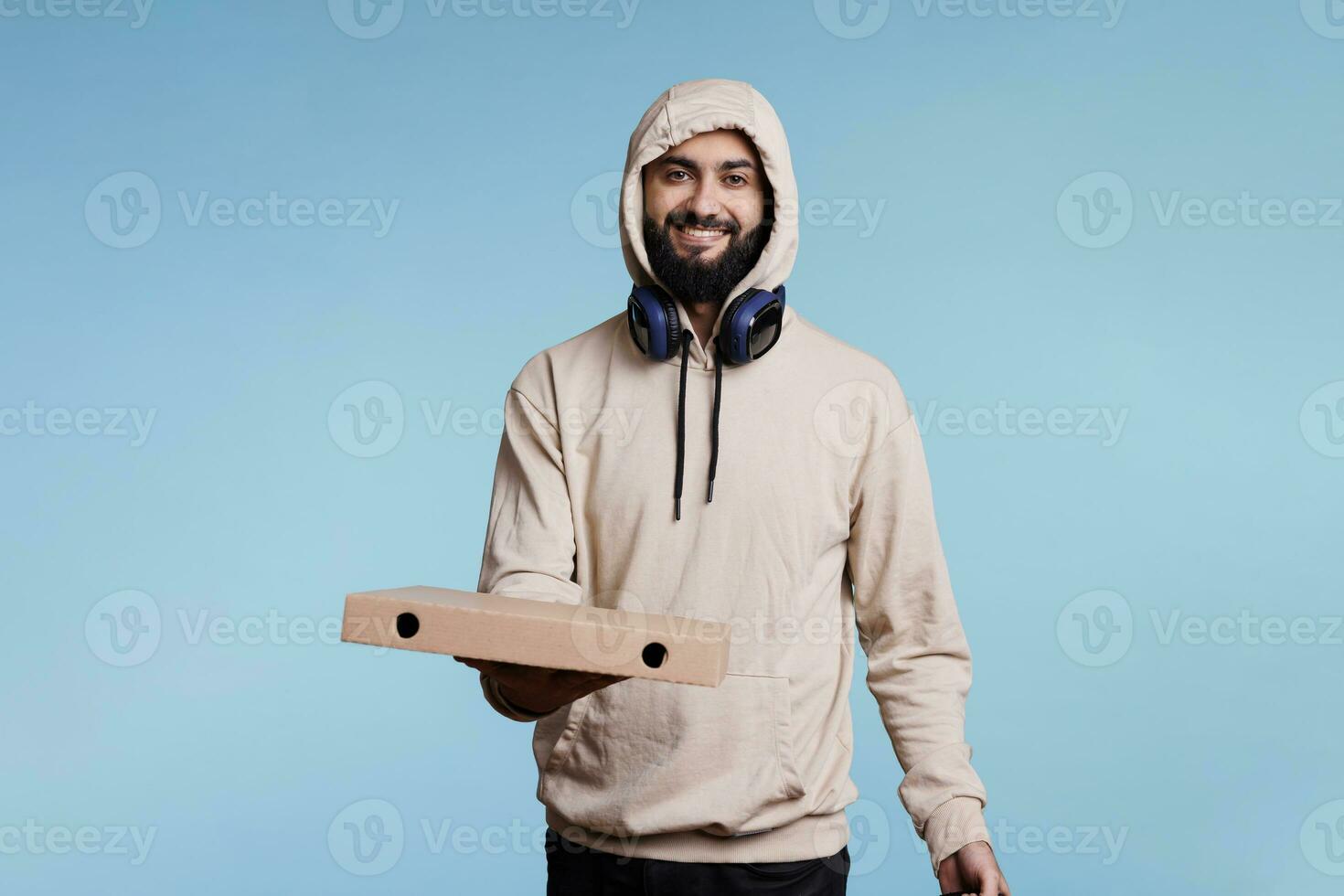 Smiling arab courier holding pizza box and looking at camera with cheerful expression. Carefree pizzeria deliveryman giving takeaway fastfood studio portrait on blue background photo