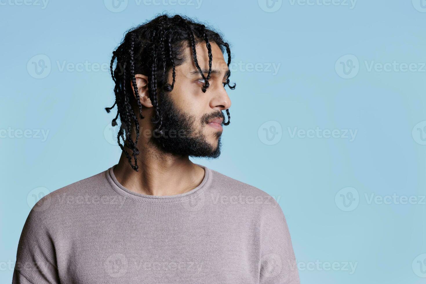 árabe hombre con neutral facial expresión mirando a izquierda lado. joven adulto árabe hermoso modelo con negro trenzado pelo posando para perfil estudio Disparo en azul antecedentes foto