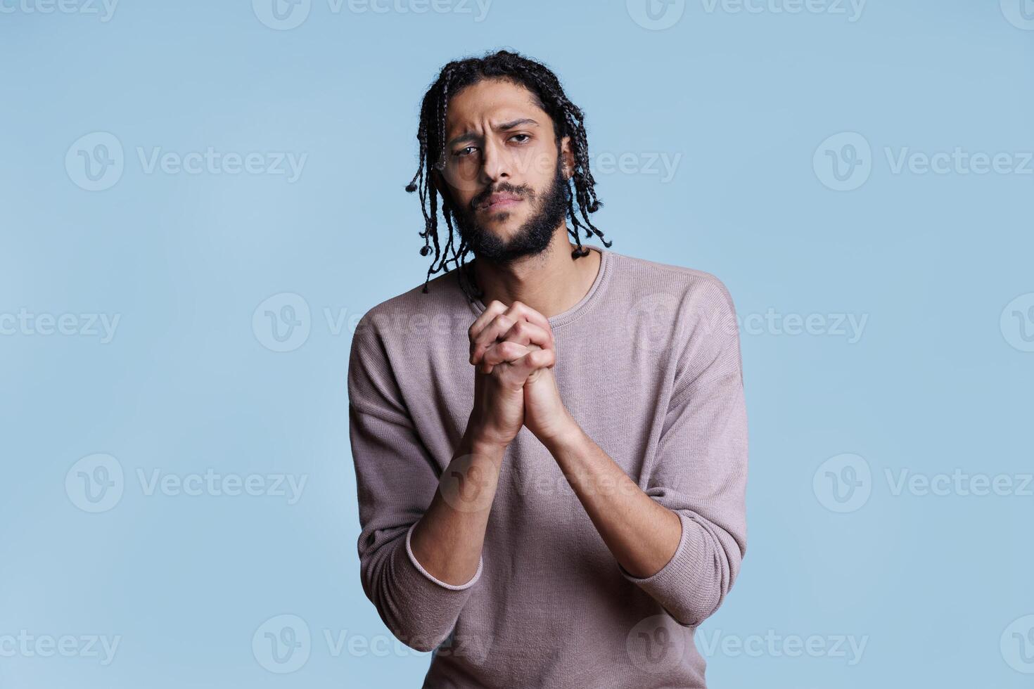 Arab man with clenched hands looking at camera with pleading facial expression. Person praying with folded arms gesture while asking for forgiveness with hopeful emotions photo