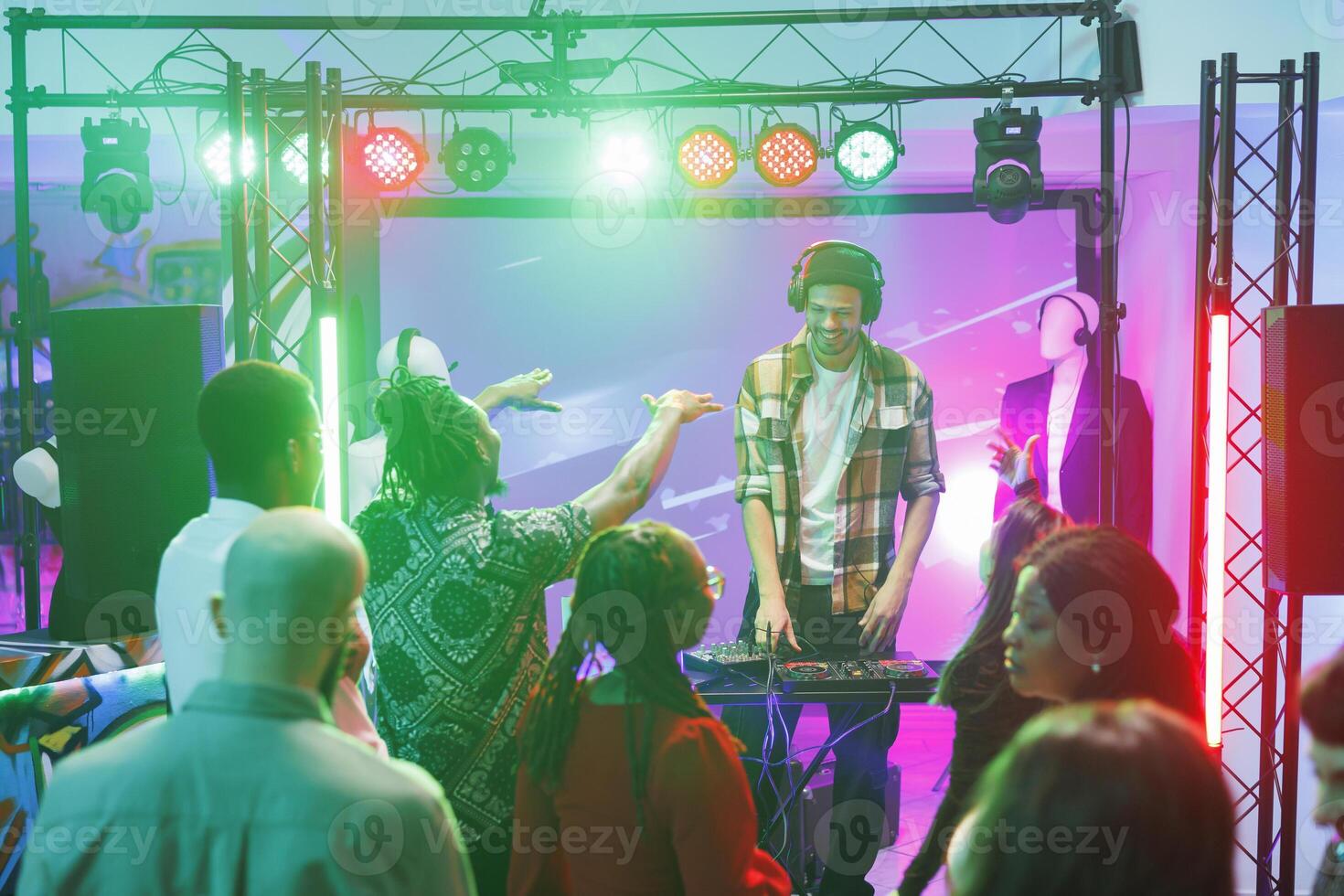 People partying on dancefloor and enjoying nightlife entertainment at discotheque in nightclub. Crowd dancing while dj performing and mixing electronic music on stage in nightclub photo