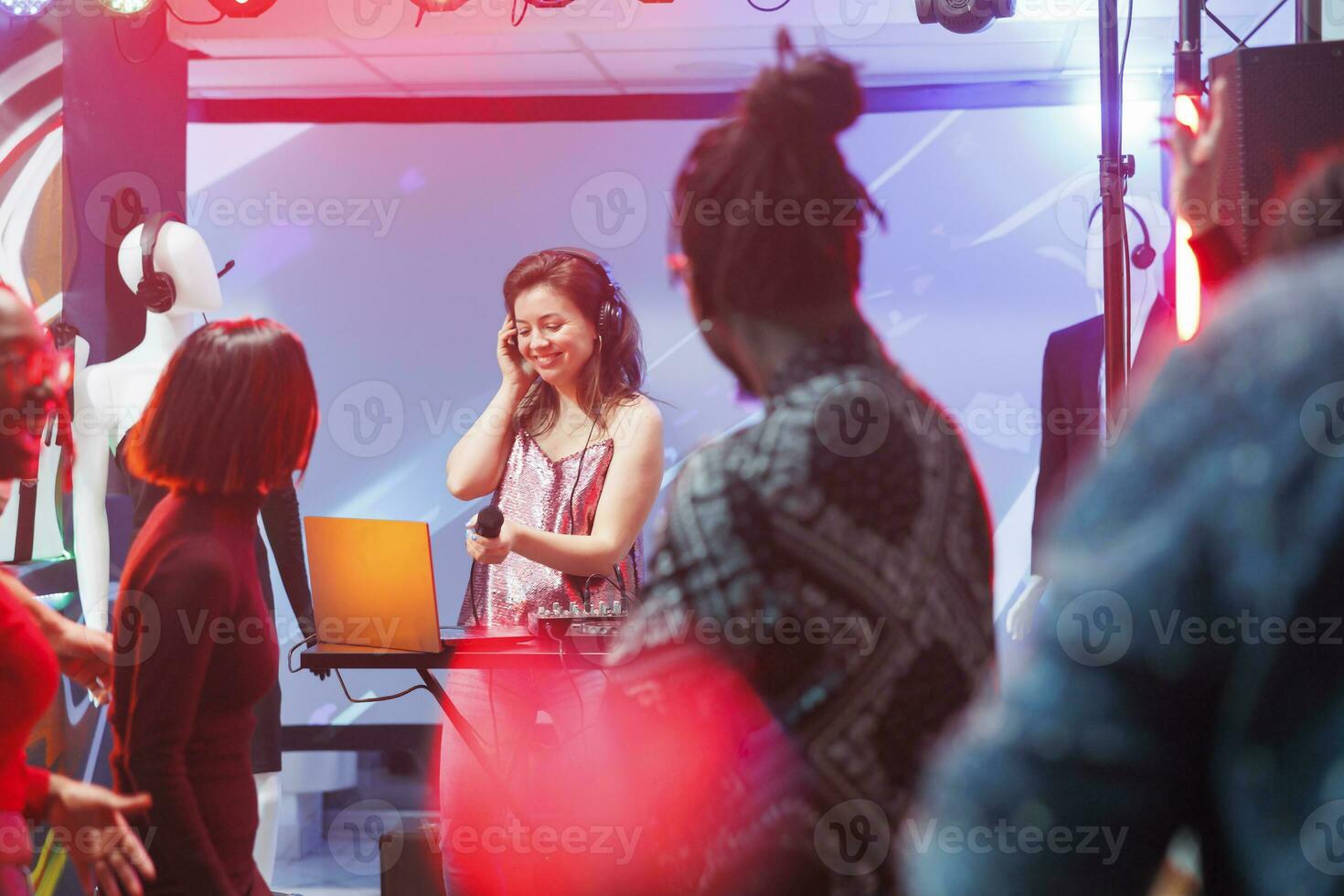 Smiling woman dj performing on stage while people dancing in nightclub at live concert. Young musician singing in microphone and mixing music at discotheque party in club photo