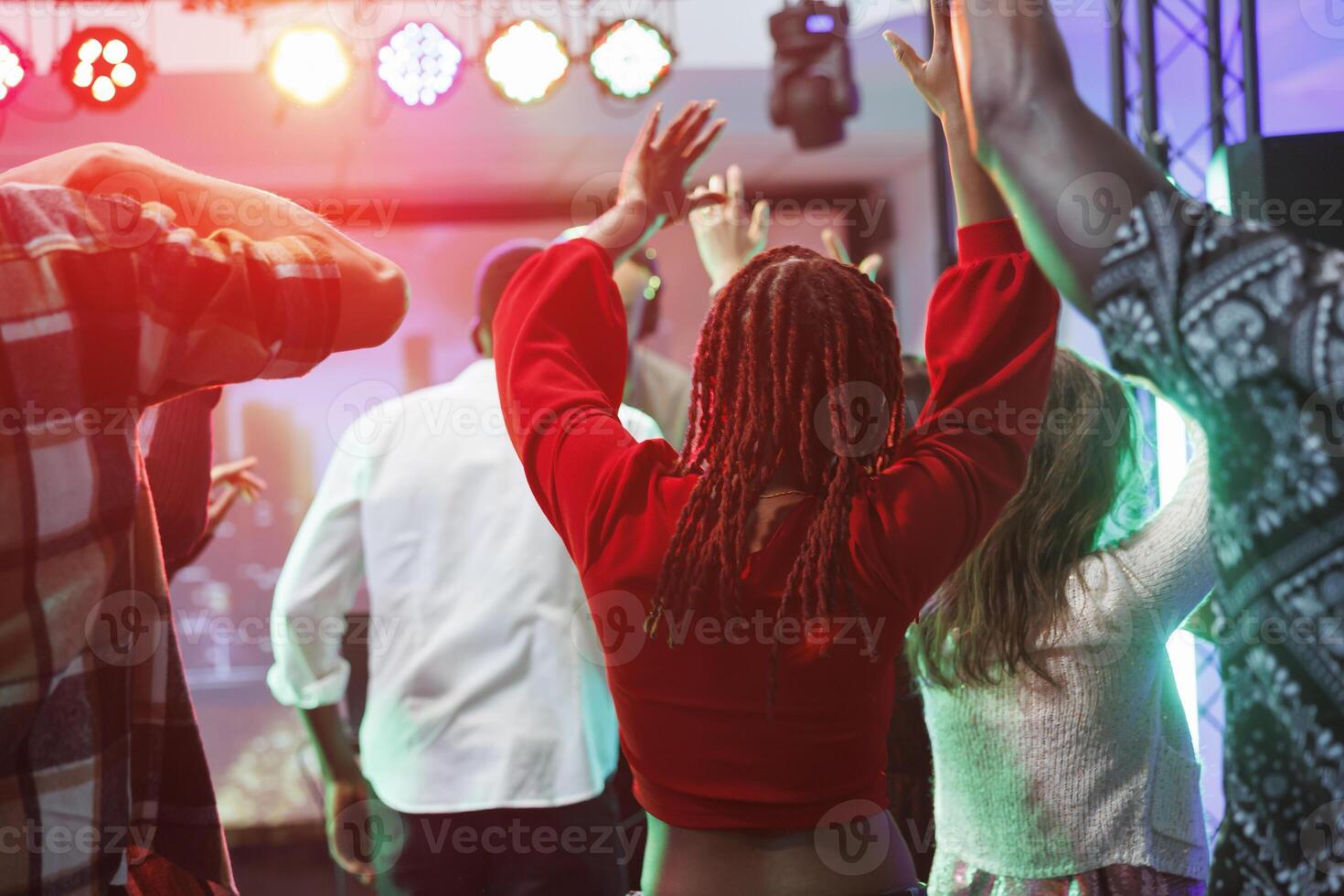 People dancing and raising hands while attending dj party in nightclub. Clubbers having fan and jumping on crowded dancefloor at discotheque in club illuminated with vibrant lights photo