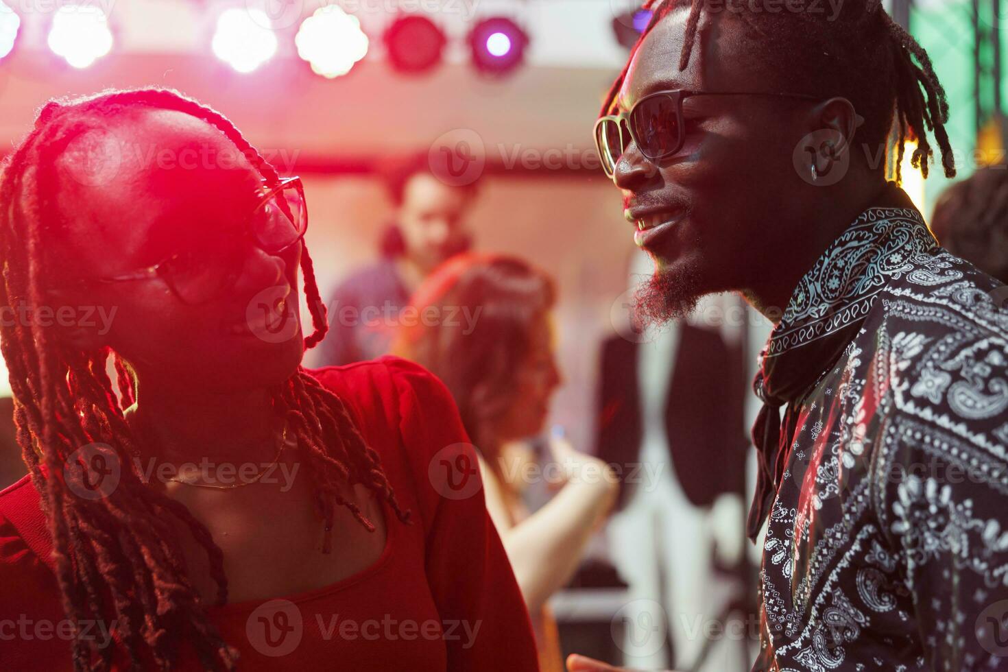 Smiling african american couple laughing and chatting in dark nightclub illuminated with vibrant lights. Carefree man and woman talking and socializing while partying in club photo