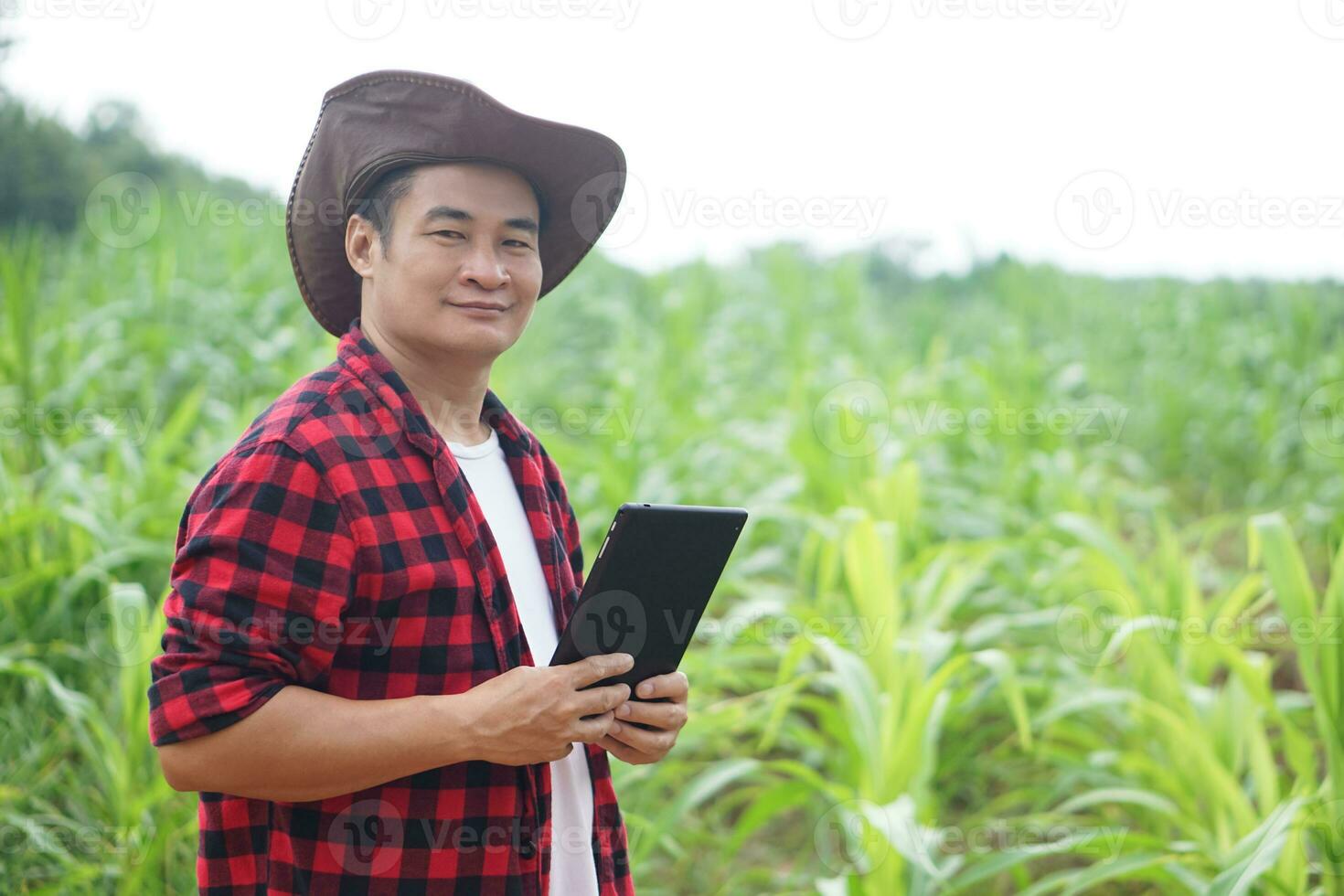 Handsome Asian man farmer is in garden, wears hat, red plaid shirt, holds  smart tablet. Feel confident, point finger, proud in crops. Concept, Agriculture occupation, inspect, survey and research. photo