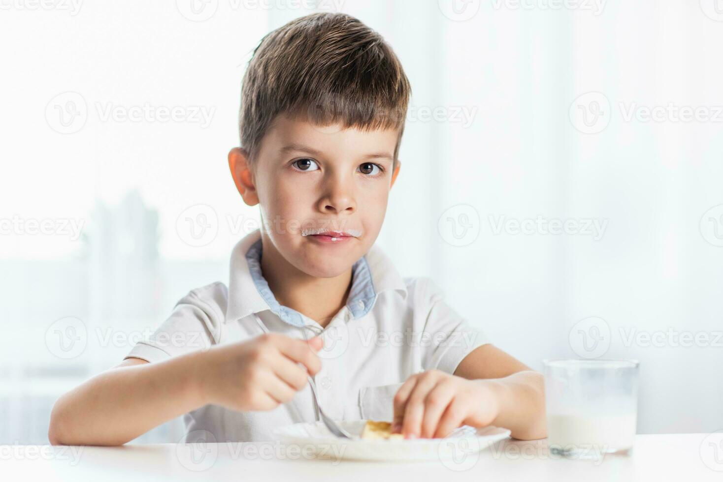 alegre chico en blanco camisa come cabaña queso tarta y bebidas Leche para desayuno a hogar foto