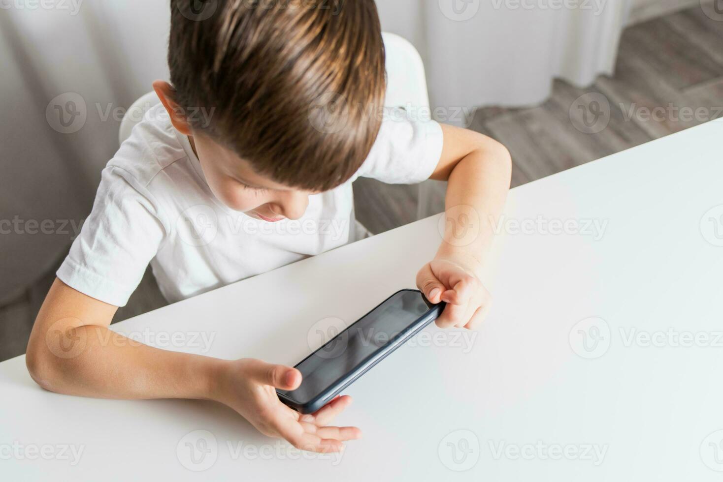 A little boy in a white T-shirt plays games on the phone at home. A happy child looks at his smartphone. photo