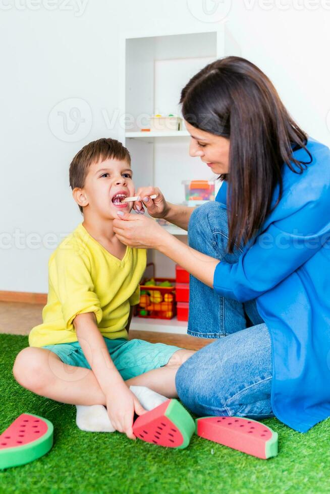 A woman speech therapist deals with a boy and performs an exercise to correct the speech apparatus by playing on the floor photo