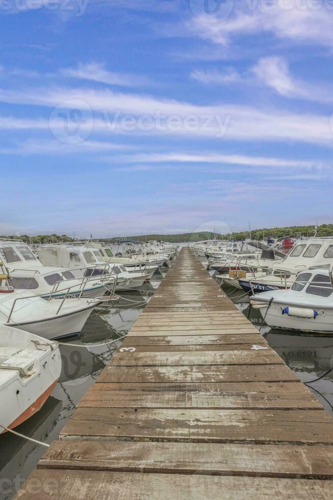 View along a wooden jetty in a harbor with docked ships in front of blue skies photo