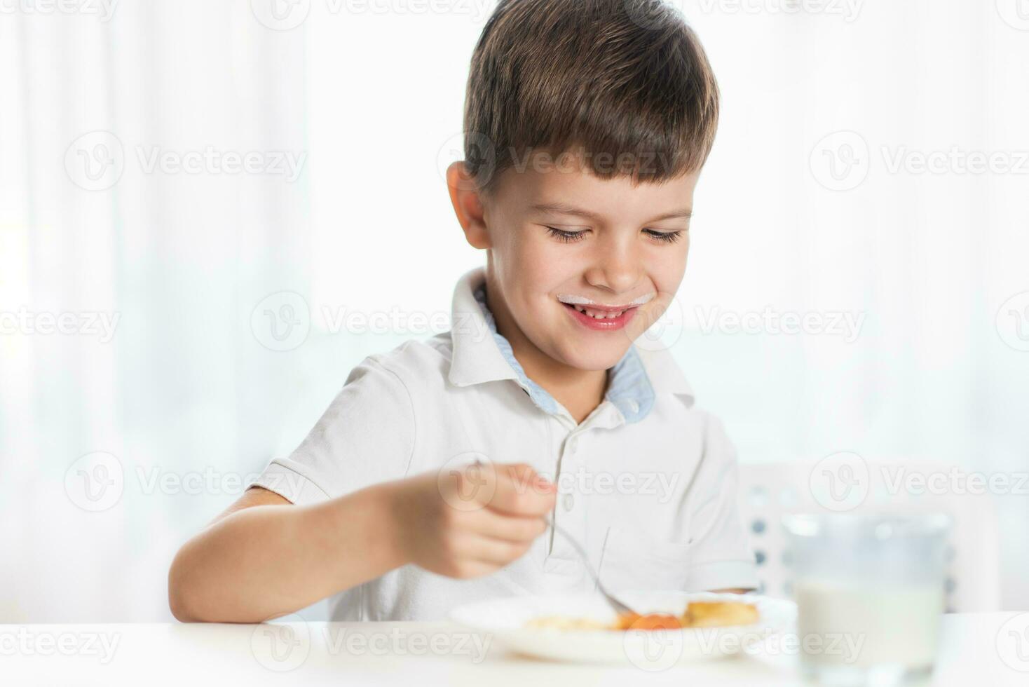 alegre chico en blanco camisa come cabaña queso tarta y bebidas Leche para desayuno a hogar foto