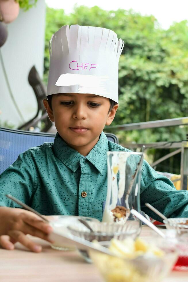 Cute Indian chef boy preparing sundae dish as a part of non fire cooking which includes vanilla ice cream, brownie, coco powder, freshly chopped fruits and strawberry syrup. Little kid preparing food photo