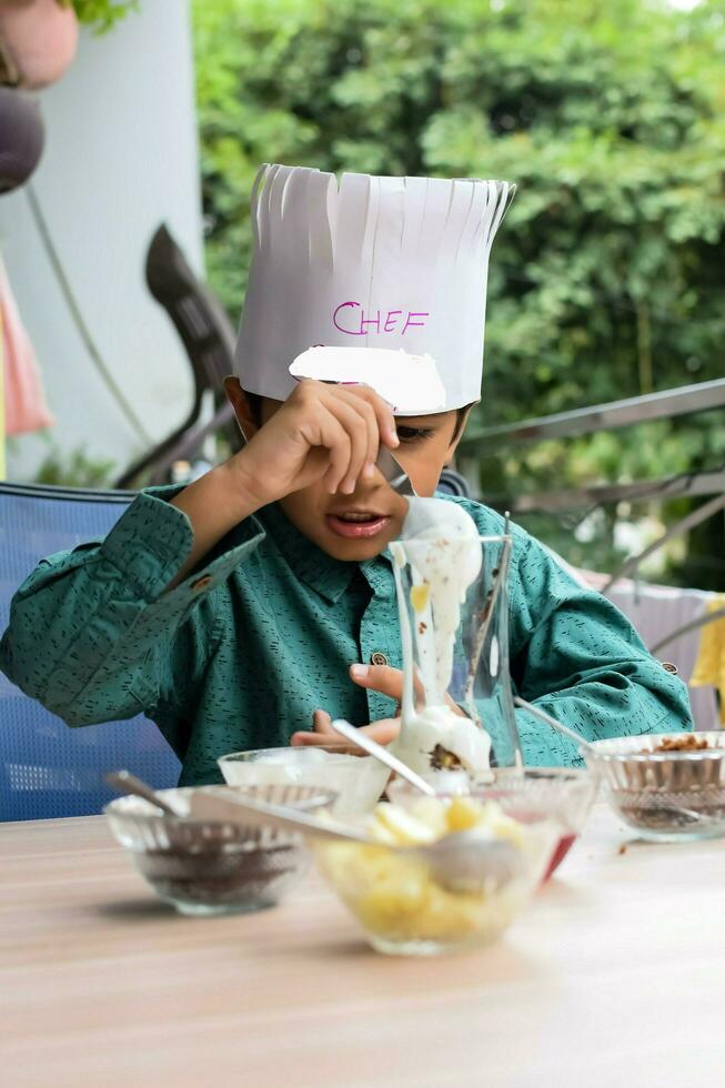 Cute Indian chef boy preparing sundae dish as a part of non fire cooking which includes vanilla ice cream, brownie, coco powder, freshly chopped fruits and strawberry syrup. Little kid preparing food photo