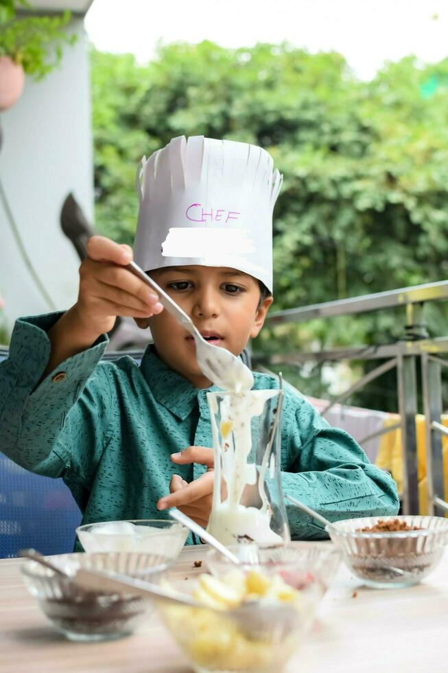 Cute Indian chef boy preparing sundae dish as a part of non fire cooking which includes vanilla ice cream, brownie, coco powder, freshly chopped fruits and strawberry syrup. Little kid preparing food photo