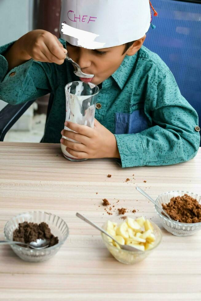 Cute Indian chef boy preparing sundae dish as a part of non fire cooking which includes vanilla ice cream, brownie, coco powder, freshly chopped fruits and strawberry syrup. Little kid preparing food photo