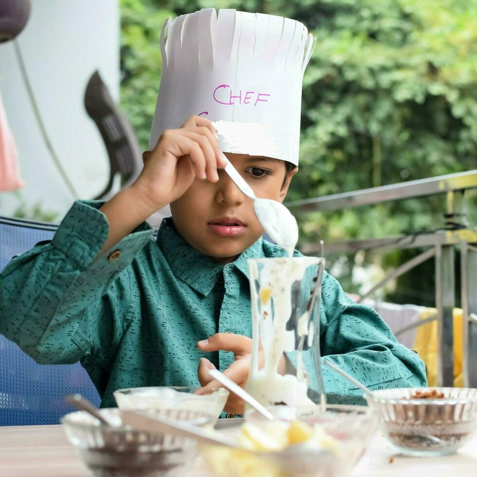 Cute Indian chef boy preparing sundae dish as a part of non fire cooking which includes vanilla ice cream, brownie, coco powder, freshly chopped fruits and strawberry syrup. Little kid preparing food photo