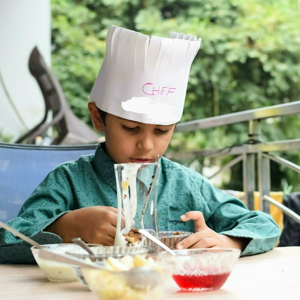 Cute Indian chef boy preparing sundae dish as a part of non fire cooking which includes vanilla ice cream, brownie, coco powder, freshly chopped fruits and strawberry syrup. Little kid preparing food photo