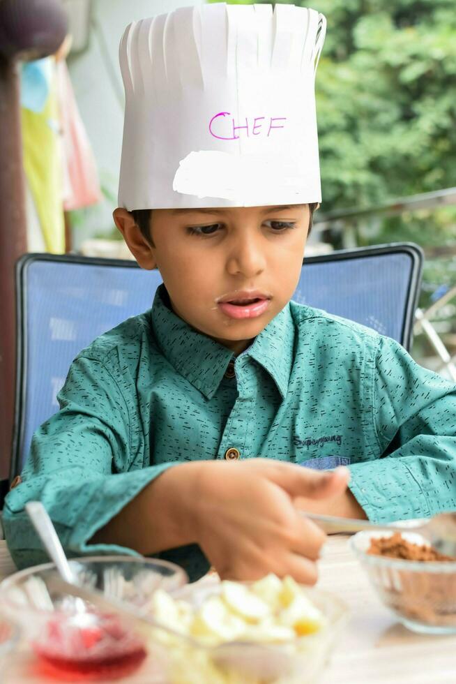 Cute Indian chef boy preparing sundae dish as a part of non fire cooking which includes vanilla ice cream, brownie, coco powder, freshly chopped fruits and strawberry syrup. Little kid preparing food photo