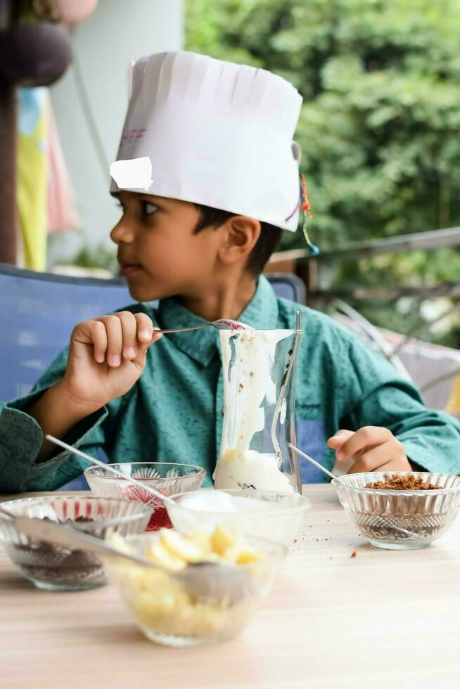 Cute Indian chef boy preparing sundae dish as a part of non fire cooking which includes vanilla ice cream, brownie, coco powder, freshly chopped fruits and strawberry syrup. Little kid preparing food photo