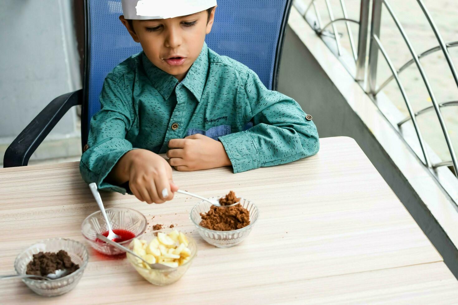 Cute Indian chef boy preparing sundae dish as a part of non fire cooking which includes vanilla ice cream, brownie, coco powder, freshly chopped fruits and strawberry syrup. Little kid preparing food photo