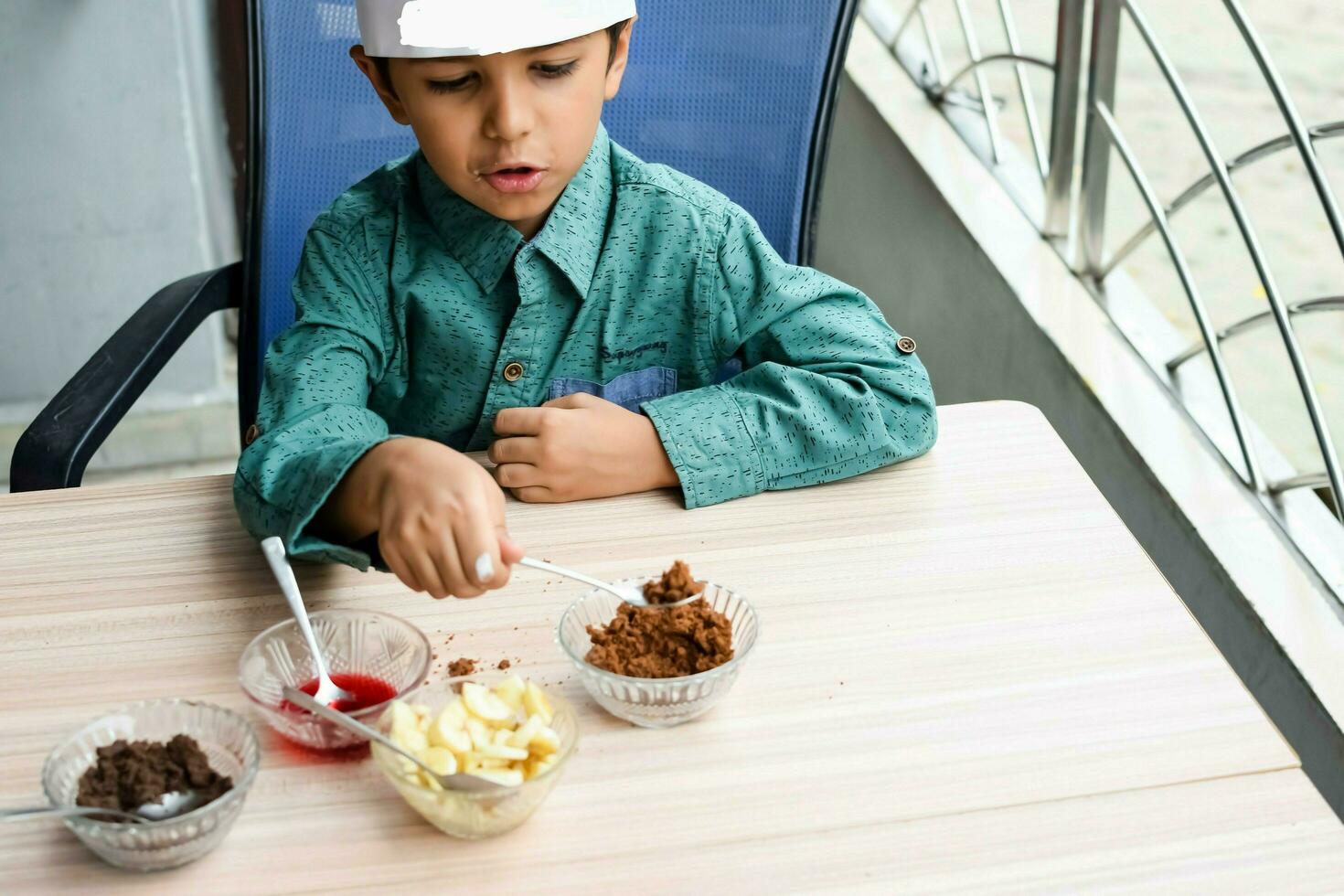 linda indio cocinero chico preparando helado con frutas y nueces plato como un parte de no fuego Cocinando cuales incluye vainilla hielo crema, duende, Coco polvo, recién Cortado frutas y fresa jarabe. pequeño niño preparando comida foto