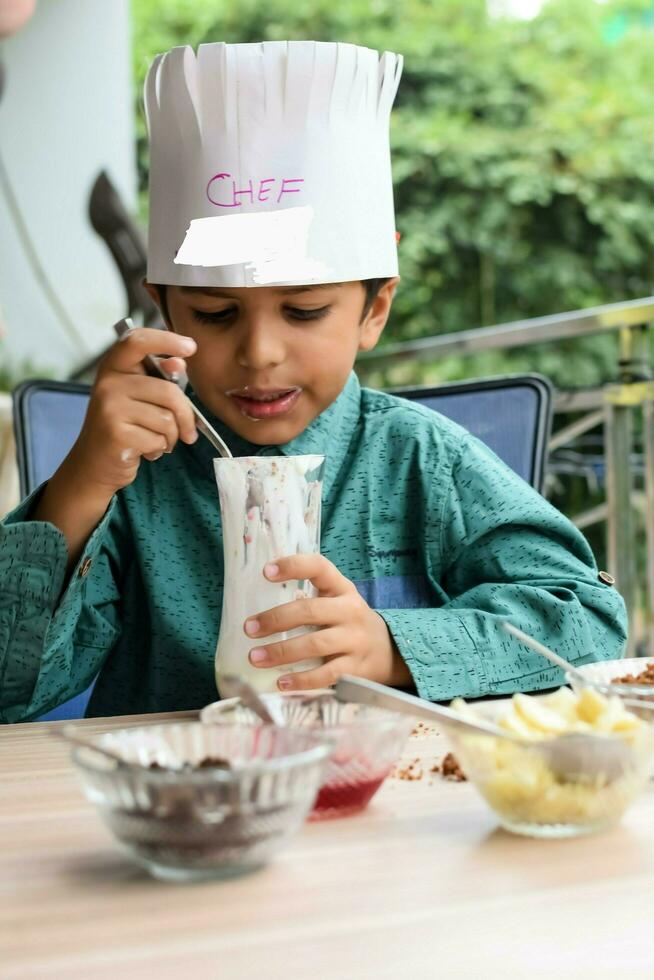 Cute Indian chef boy preparing sundae dish as a part of non fire cooking which includes vanilla ice cream, brownie, coco powder, freshly chopped fruits and strawberry syrup. Little kid preparing food photo