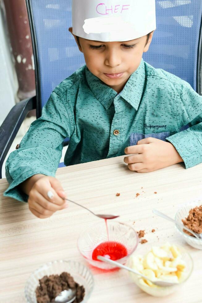 Cute Indian chef boy preparing sundae dish as a part of non fire cooking which includes vanilla ice cream, brownie, coco powder, freshly chopped fruits and strawberry syrup. Little kid preparing food photo