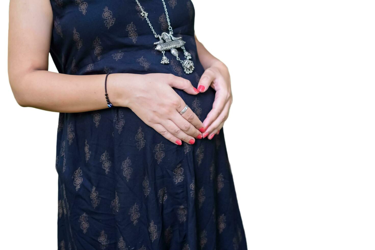 A pregnant Indian lady poses for pregnancy shoot and hands on belly with white background, Indian pregnant woman puts her hand on her stomach in maternity dress with plain background, Pregnancy shoot photo