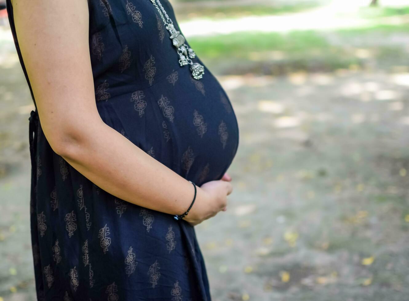 un embarazada indio dama poses para al aire libre el embarazo disparar y manos en barriga, indio embarazada mujer pone su mano en su estómago con un maternidad vestir a sociedad parque, embarazada fuera de maternidad disparar foto