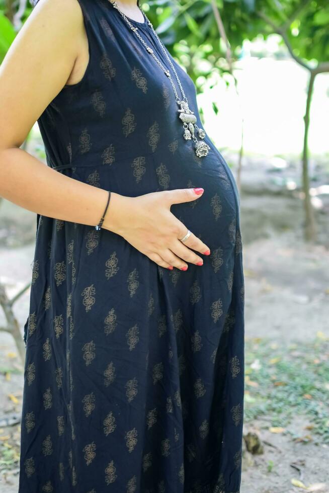 A pregnant Indian lady poses for outdoor pregnancy shoot and hands on belly, Indian pregnant woman puts her hand on her stomach with a maternity dress at society park, Pregnant outside maternity shoot photo