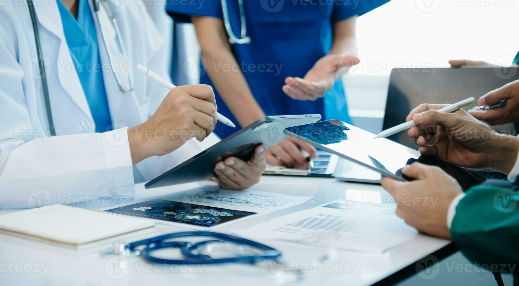 Medical team having a meeting with doctors in white lab coats and surgical scrubs seated at a table discussing a patients working online using computers photo
