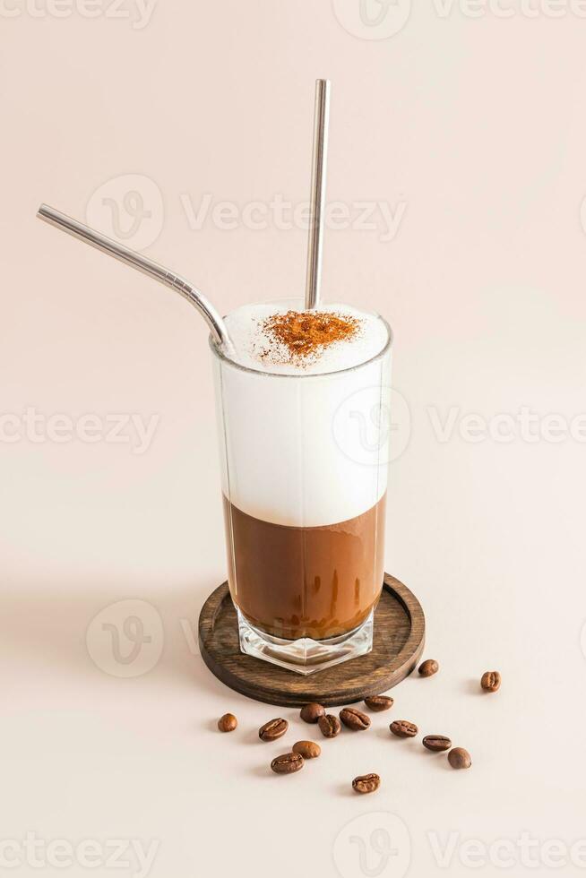 A tall glass of delicious coffee with chic milk foam with eco straw stands on a wooden plate with coffee beans. Vertical front view. Beige background. photo