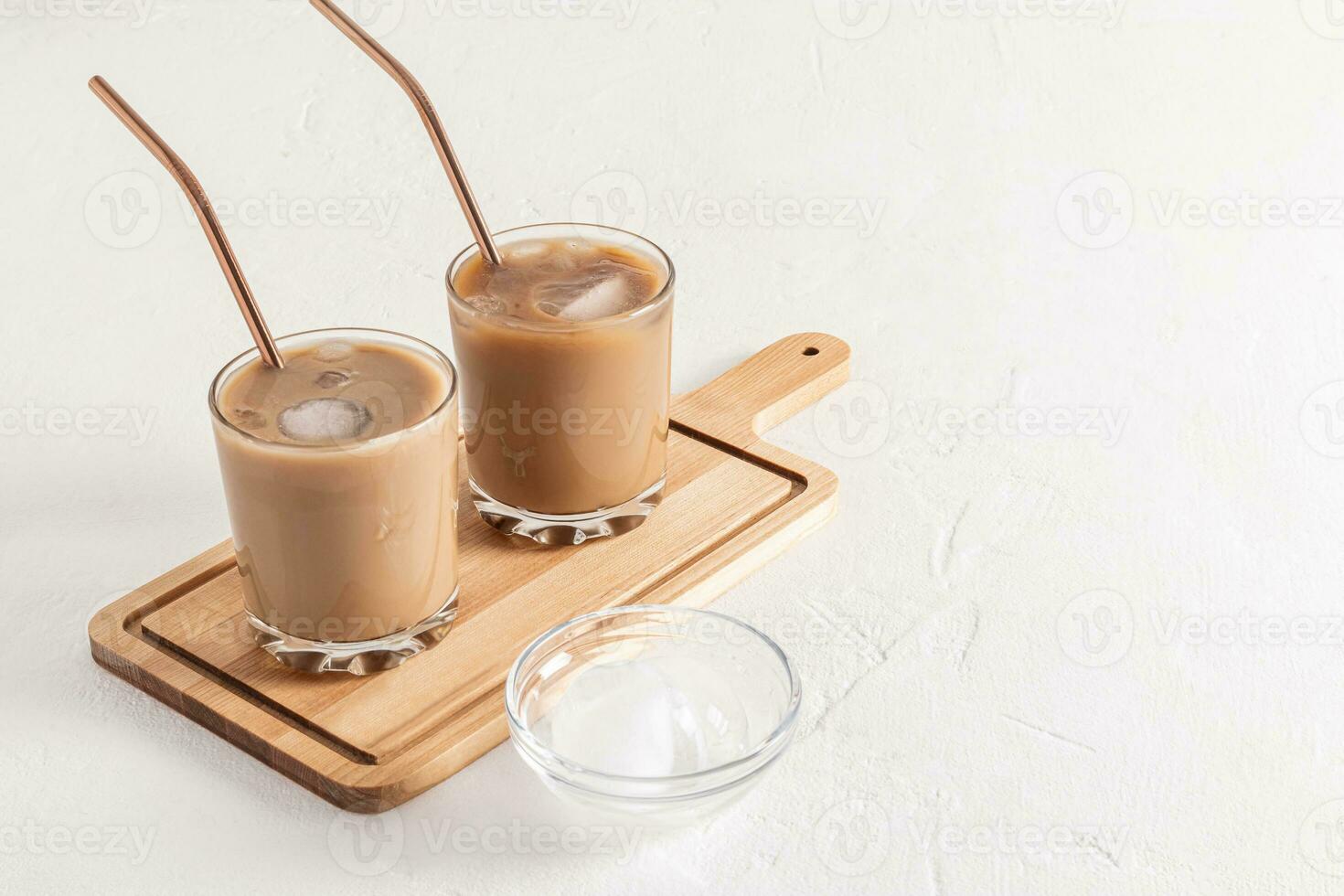 Two glasses of delicious cool dolgone coffee with ice cubes and eco straw stand on a wooden cutting board. White textured background. A copy space photo