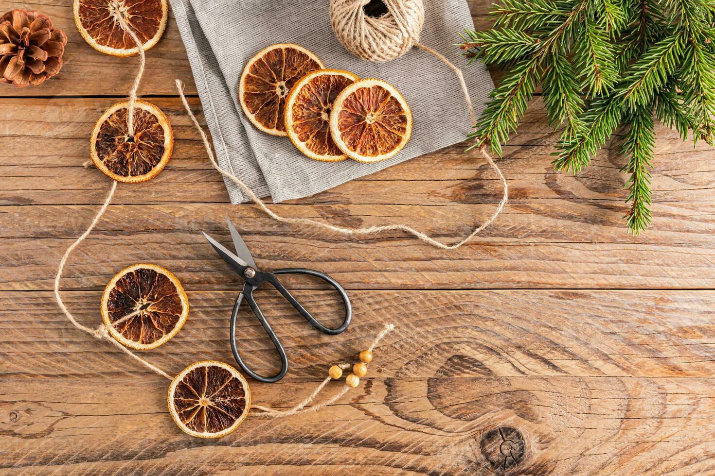 Homemade garland of dried oranges on a wooden rustic table, vintage scissors, a skein of thread for crafts, spruce branches. Top view. photo