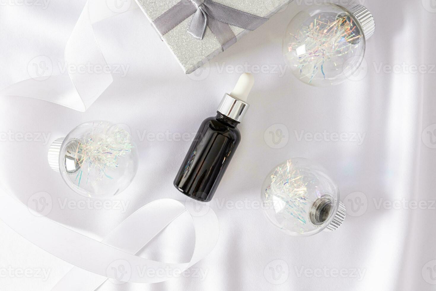 Cosmetic bottle with dropper and silver cap lies on white satin among silver Christmas balls and gift box. Top view. Empty product packaging. photo