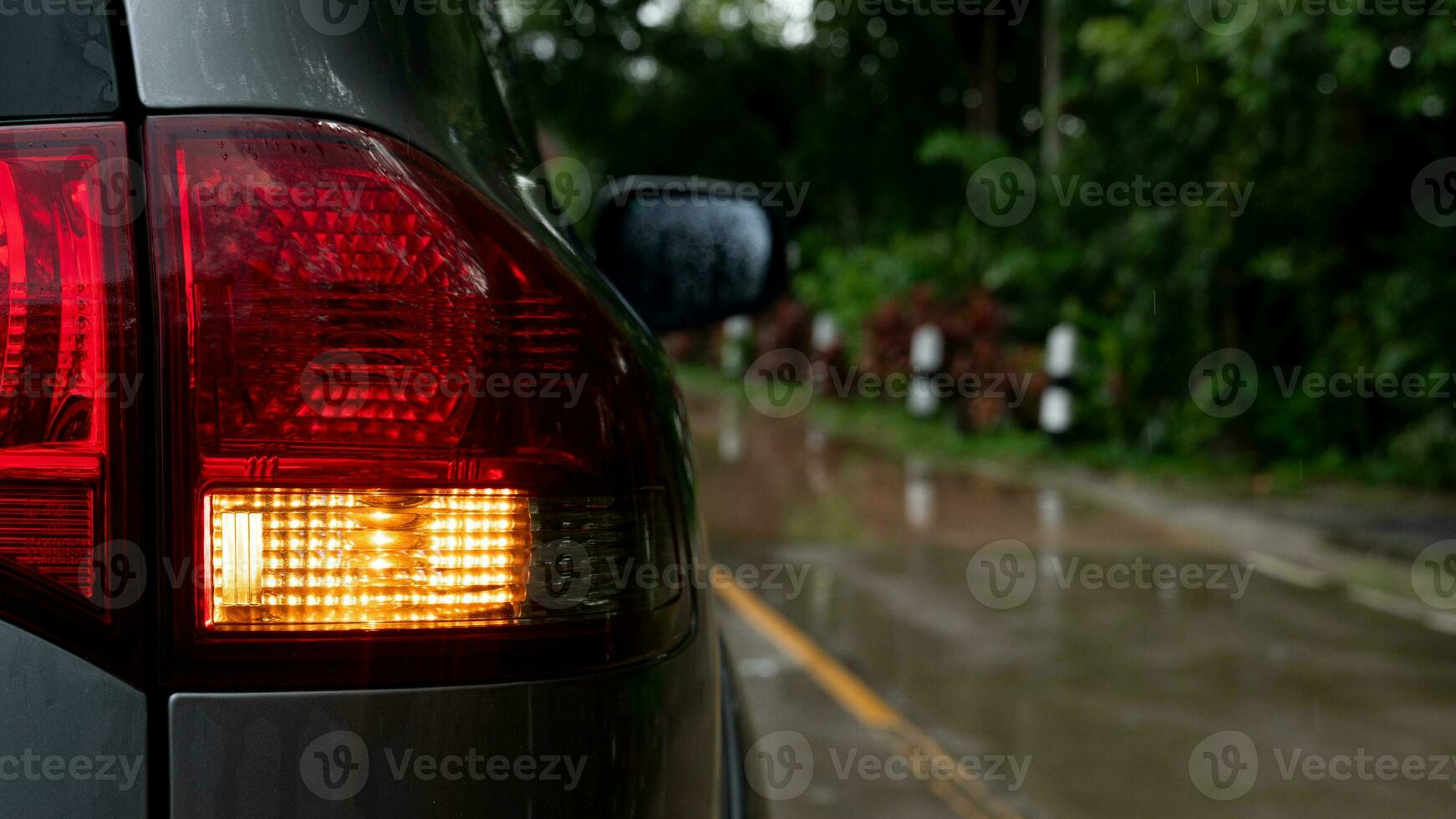 Close-up of turn light signal of gray car with water of rain on surface. Driving on wet cement road from rainy. Beside road with green forest. photo